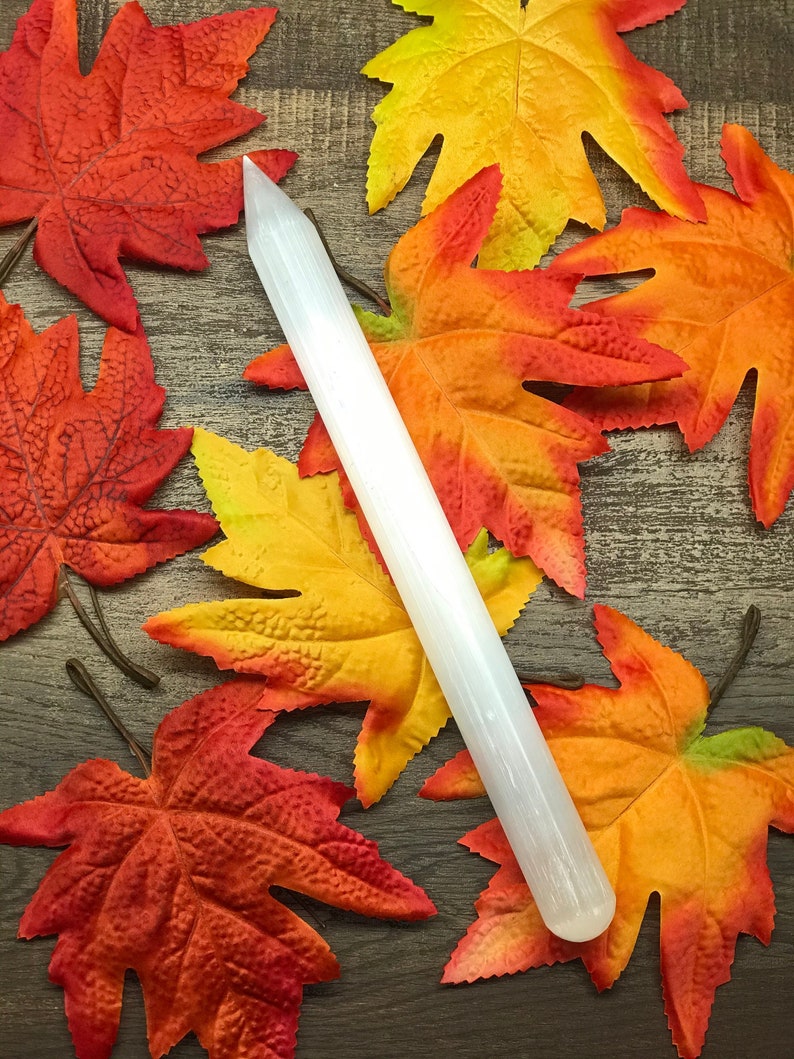 Puede incluir: Una varita de selenita blanca se encuentra en diagonal sobre una superficie de madera, rodeada de coloridas hojas de oto&ntilde;o en tonos rojos, naranjas y amarillos. La varita es cil&iacute;ndrica con una punta. Las hojas a&ntilde;aden un toque estacional.