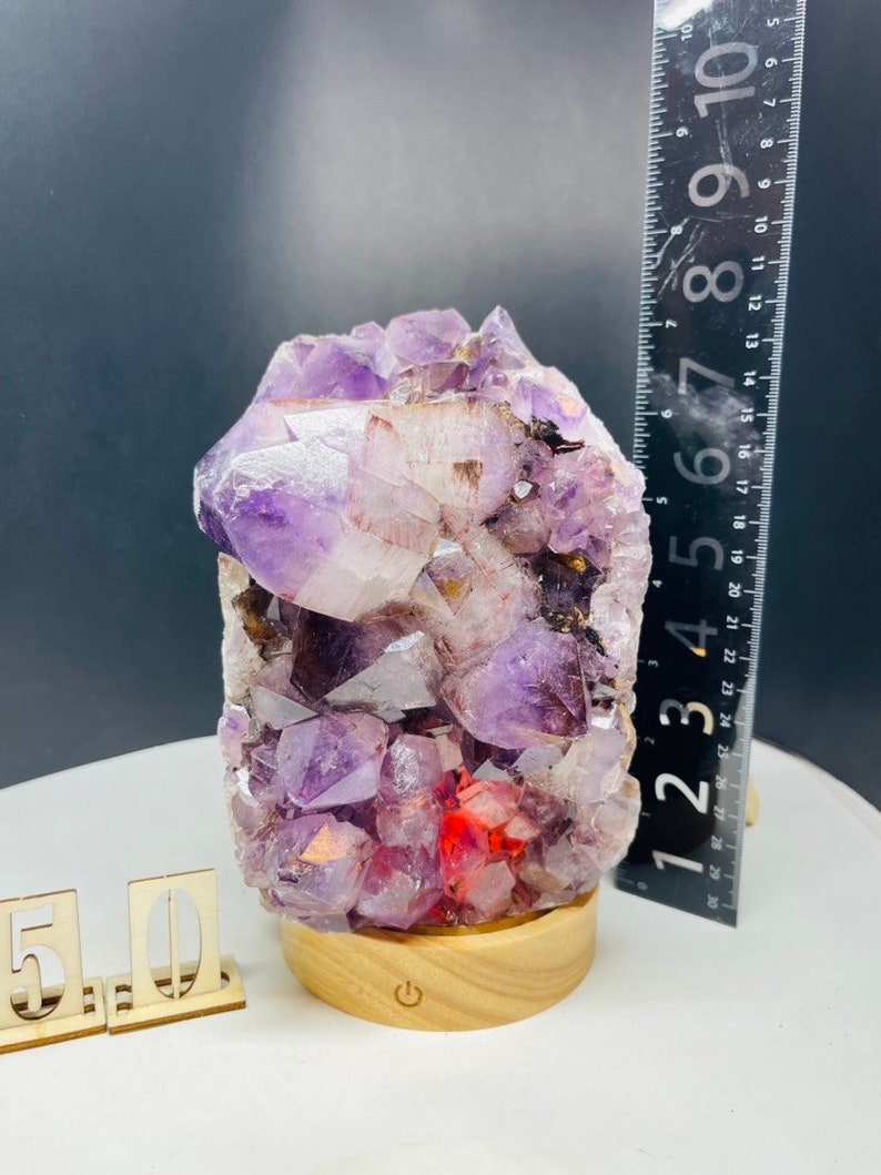 May include: A purple amethyst crystal cluster with red inclusions, mounted on a wooden base with a light switch. The crystal is approximately 7 inches tall.