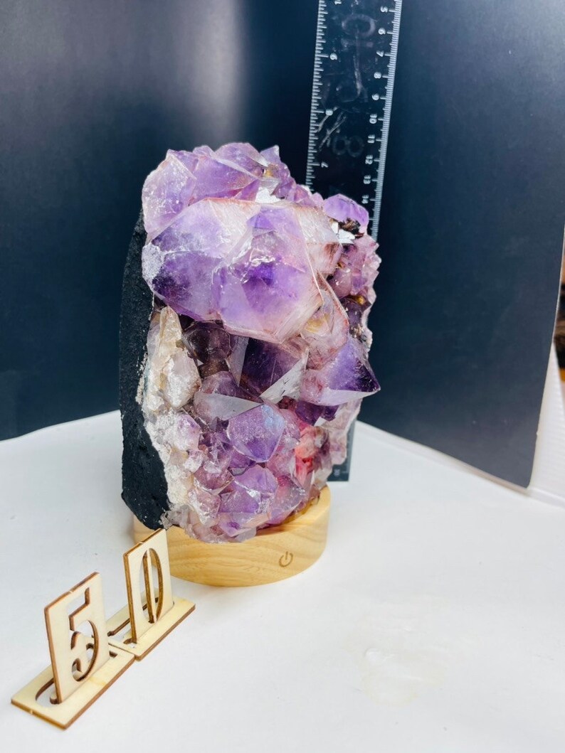 May include: A large amethyst crystal cluster on a wooden base with a light switch. The crystal is purple and has a natural, rough texture. The base is made of wood and has a natural finish.