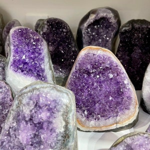 May include: A collection of amethyst crystal geodes, each with a unique shape and size. The crystals are a deep purple colour and have a natural, rough texture. The geodes are displayed on a white surface.