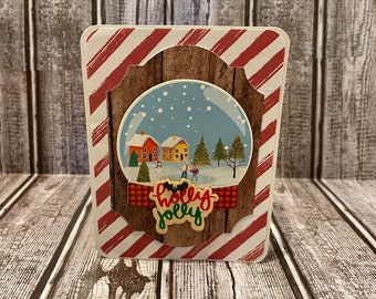 May include: A Christmas card with a red and white striped background. The card features a snow globe with a winter scene inside. The snow globe is surrounded by a wooden frame and a red and black plaid ribbon. The text "holly jolly" is written in gold letters on the card.