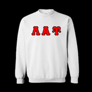 May include: A white crewneck sweatshirt featuring the Greek letters Lambda, Alpha, and Tau printed in red with a black outline across the chest. The garment has long sleeves, a ribbed collar, and ribbed cuffs, providing a standard fit and design.