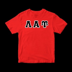May include: A bright red short-sleeved t-shirt featuring the Greek letters Lambda, Alpha, and Tau printed across the chest. The letters are black with a thick white outline, creating a bold contrast against the solid red fabric of the shirt.