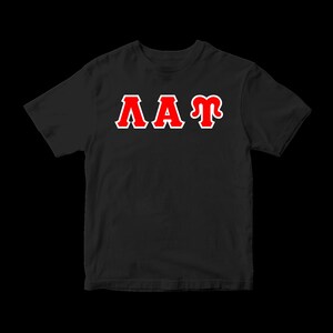 May include: A black short-sleeved t-shirt featuring the Greek letters Lambda, Alpha, and Tau printed across the chest. The letters are rendered in a bold red color with a distinct white outline, creating a high-contrast graphic design on the dark fabric.