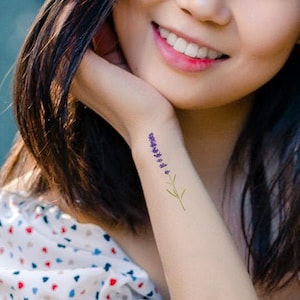 May include: A woman with long dark hair is wearing a white dress with a red and blue heart pattern. She has a small lavender flower tattoo on her wrist.