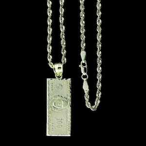May include: A gold chain necklace with a gold pendant shaped like a $100 bill. The pendant has a detailed design and features the portrait of Benjamin Franklin.