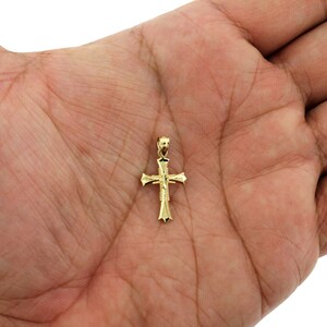 May include: A small gold cross pendant with a detailed design. The cross is on a hand, showing its size.