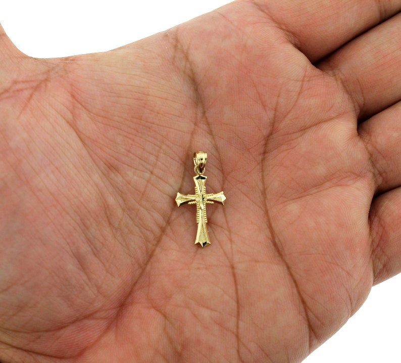 May include: A small gold cross pendant with a detailed design. The cross is on a hand, showing its size.