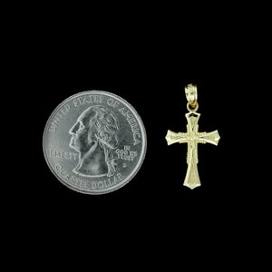 May include: A gold-colored cross pendant, shown next to a US quarter for size comparison. The cross has a simple design with a slightly rounded edge.
