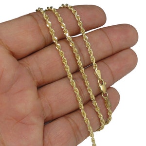 May include: A gold rope chain necklace with a lobster clasp closure. The chain is shown on a hand, with the chain laying in a loose, folded pattern.