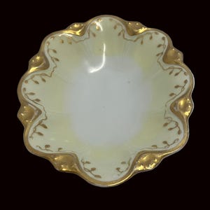 May include: A small, scalloped-edge dish with a pale yellow interior and a white center. The rim and edges are trimmed with gold, and gold leaf designs are visible. The dish is likely made of porcelain or ceramic.