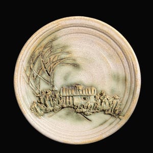 May include: A round, off-white ceramic plate with a raised relief design. The design features a tree, a building, and other architectural elements. The plate has a textured surface and a dark background.
