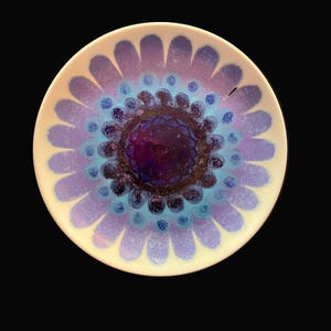May include: A round ceramic plate featuring a vibrant floral pattern with a deep purple center, surrounded by concentric rings of dark blue, light blue, and lavender petals. The design radiates outward from the center against a white background.