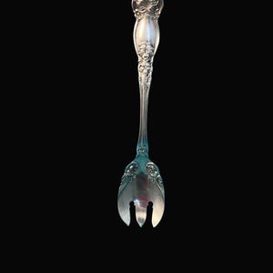 May include: A silver-toned metal serving fork featuring an ornate handle with embossed floral detailing at the top and base of the stem. The bowl of the fork has three wide tines with decorative scrollwork patterns integrated into the metal surface.
