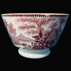 May include: A white and maroon ceramic bowl with a detailed landscape design. The bowl features a footed base and a rim with a repeating triangular pattern. The landscape includes trees, flowers, and a mountain.