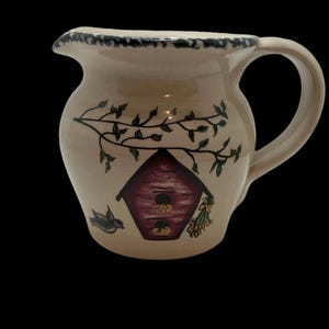 May include: A cream-colored ceramic pitcher with a birdhouse and a bird painted on it. The birdhouse is red with a brown roof and has two holes for birds to enter. The bird is blue and white and is flying towards the birdhouse. There are green leaves and yellow flowers painted on the pitcher.