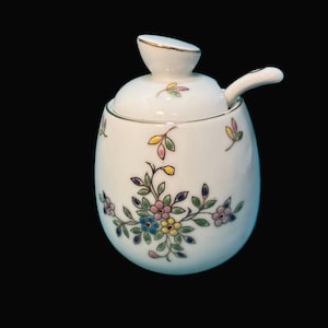 May include: White ceramic sugar bowl with lid and spoon. The bowl is decorated with a floral design in pink, blue, yellow, and green. The rim and handle are trimmed with gold.