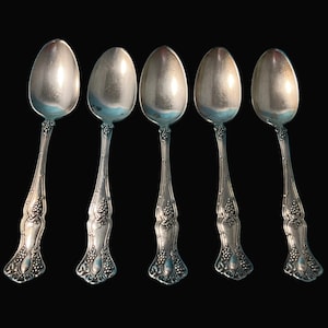 May include: Five silver spoons with ornate handles and oval bowls are arranged against a black background. The handles feature a detailed floral and grape design. The spoons are likely for serving or dining.