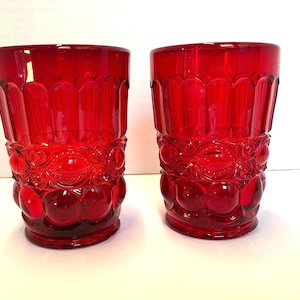 May include: Two ruby red drinking glasses. Each glass has a detailed pattern of vertical lines, circular designs, and rounded shapes. The glasses are made of a transparent material, allowing light to pass through.