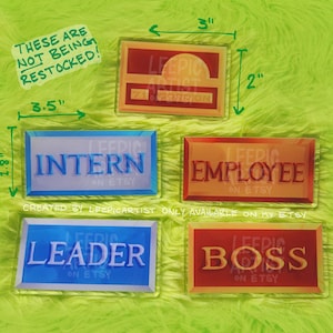 Może przedstawiać: Cztery akrylowe odznaki z różnymi tytułami: Stażysta, Pracownik, Lider i Szef. Każda odznaka ma 7,6 cm szerokości i 4,6 cm wysokości. Odznaki znajdują się na zielonej, puszystej powierzchni. Tekst na odznakach jest biały, a kolory tła to niebieski, pomarańczowy, niebieski i czerwony.