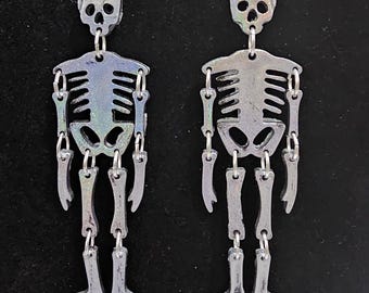 May include: Two silver-colored skeleton earrings. Each earring features a skull, rib cage, and articulated limbs. The earrings are suspended from silver-colored fish hook ear wires. The skeletons have a reflective, iridescent finish, adding a touch of sparkle.