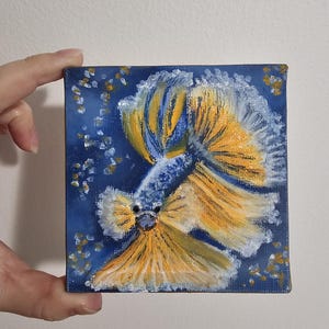 May include: Square canvas painting of a Betta fish. The fish has a blue body and yellow fins with white accents. The background is a dark blue with small white and gold dots.