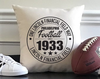 May include: A white throw pillow with a black circle design that says "One Lincoln Financial Field Way. Philadelphia Football Since 1933. Lincoln Financial Field." A brown football with white stitching and the NFL logo is on the right side of the pillow. Two white trainers with black stripes are on either side of the pillow. The pillow, football, and trainers are on a black shelf with two wicker baskets underneath.