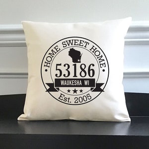 May include: A white throw pillow with a black circle design that says "Home Sweet Home" with a silhouette of the state of Wisconsin, the zip code 53186, the text "Waukesha WI", three stars, and "Est. 2005". The pillow is sitting on a black bench with two wicker baskets underneath.