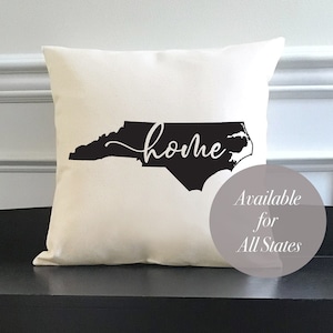 May include: A white throw pillow with a black outline of the state of North Carolina and the word "home" written in cursive. The pillow is sitting on a black bench with two wicker baskets underneath. A white circle with the text "Available for All States" is in the lower right corner of the image.