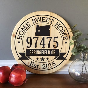 May include: A round, light-colored wooden sign featuring black text and graphics. The text reads HOME SWEET HOME, 97475, SPRINGFIELD OR, and Est. 2015. A black silhouette of the state of Oregon with a star and three black stars are also printed.