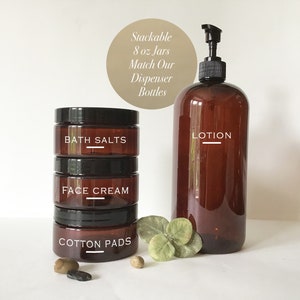 May include: Three amber glass jars with black lids stacked on top of each other. The jars are labeled "Bath Salts", "Face Cream", and "Cotton Pads". A larger amber glass bottle with a black pump top is labeled "LOTION". The text "Stackable 8 oz Jars Match Our Dispenser Bottles" is written in brown text on a tan circle.