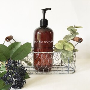 May include: A brown glass bottle with a black pump dispenser filled with hand soap. The bottle is labeled "HAND SOAP" in white text and is sitting in a silver wire basket with a wooden handle.