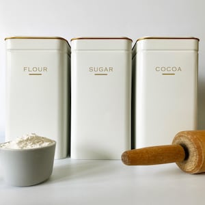 Op de afbeelding: Drie witte rechthoekige keukencontainers met de opschriften "FLOUR", "SUGAR" en "COCOA" en gouden randen. Een maatbeker met bloem en een houten deegroller staan op de voorgrond. Voor bakbenodigdheden.