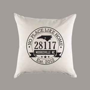 May include: White throw pillow with a black circle design that says "No Place Like Home" with a North Carolina state outline, the zip code 28117, the words "Mooresville NC", three stars, and "Est. 2012".