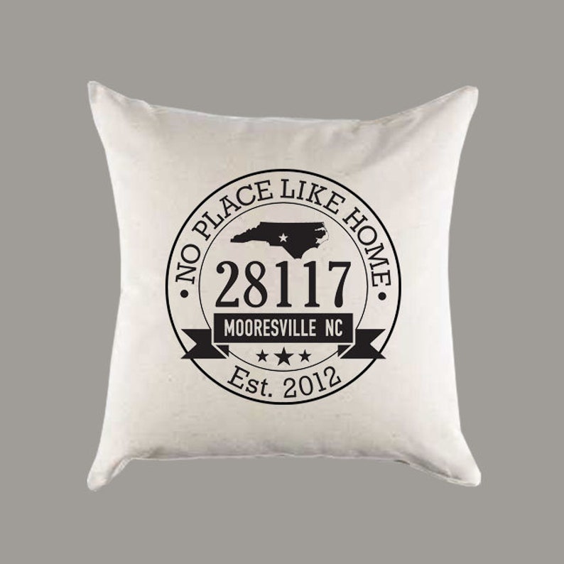 May include: White throw pillow with a black circle design that says "No Place Like Home" with a North Carolina state outline, the zip code 28117, the words "Mooresville NC", three stars, and "Est. 2012".