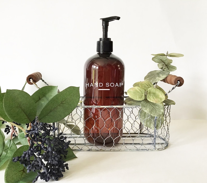 May include: A brown glass bottle with a black pump dispenser filled with hand soap. The bottle is labeled "HAND SOAP" in white text and is sitting in a silver wire basket with a wooden handle.