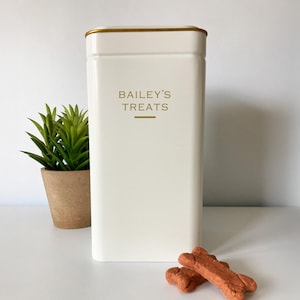 Puede incluir: Un recipiente rectangular blanco roto para golosinas para mascotas con ribete dorado y las palabras "BAILEY'S TREATS" en letras doradas. Dos golosinas para perros en forma de hueso están frente al recipiente.