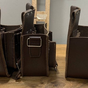 May include: Three brown leather tool bags with multiple pockets and a strap. The bags are lined up on a wooden surface.