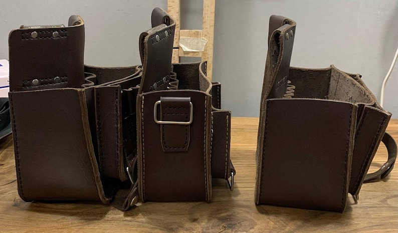 May include: Three brown leather tool bags with multiple pockets and a strap. The bags are lined up on a wooden surface.