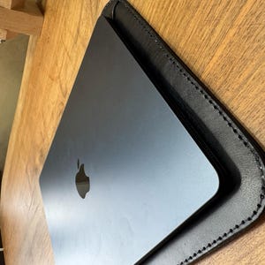 Puede incluir: Un ordenador portátil negro en una funda de cuero negro con bordes cosidos. El portátil tiene un logotipo de Apple plateado. La funda está sobre una superficie de madera.