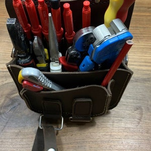 May include: A brown leather tool bag with a variety of tools, including screwdrivers, pliers, wire strippers, and a pencil. The bag has a strap and a buckle.