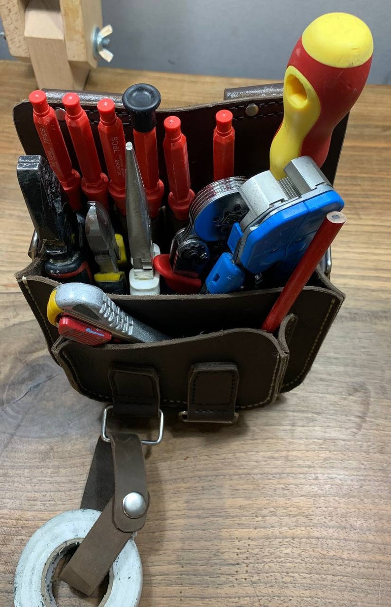 May include: A brown leather tool bag with a variety of tools, including screwdrivers, pliers, wire strippers, and a pencil. The bag has a strap and a buckle.