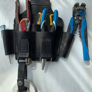Peut inclure: Une ceinture &agrave; outils en cuir noir avec plusieurs poches, contenant divers outils tels que des pinces coupantes, des pinces et un couteau utilitaire. La ceinture a une boucle pour accrocher et un rouleau de ruban adh&eacute;sif &eacute;lectrique.