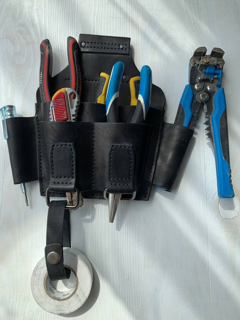 Peut inclure: Une ceinture &agrave; outils en cuir noir avec plusieurs poches, contenant divers outils tels que des pinces coupantes, des pinces et un couteau utilitaire. La ceinture a une boucle pour accrocher et un rouleau de ruban adh&eacute;sif &eacute;lectrique.