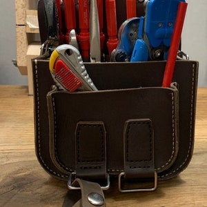May include: A brown leather tool bag with a variety of tools, including screwdrivers, wire strippers, a utility knife, and a tape measure. The bag has a strap and a buckle closure. The bag is labeled "V2".