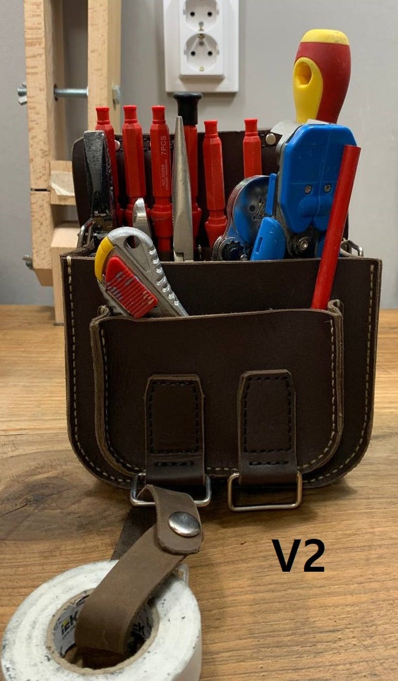 May include: A brown leather tool bag with a variety of tools, including screwdrivers, wire strippers, a utility knife, and a tape measure. The bag has a strap and a buckle closure. The bag is labeled "V2".