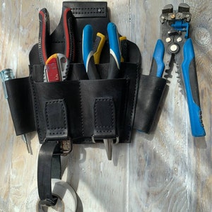 Peut inclure: Une ceinture &agrave; outils en cuir noir avec plusieurs poches, contenant divers outils tels que des pinces, des d&eacute;nudeurs de fils et un tournevis. La ceinture est accroch&eacute;e &agrave; une surface en bois.
