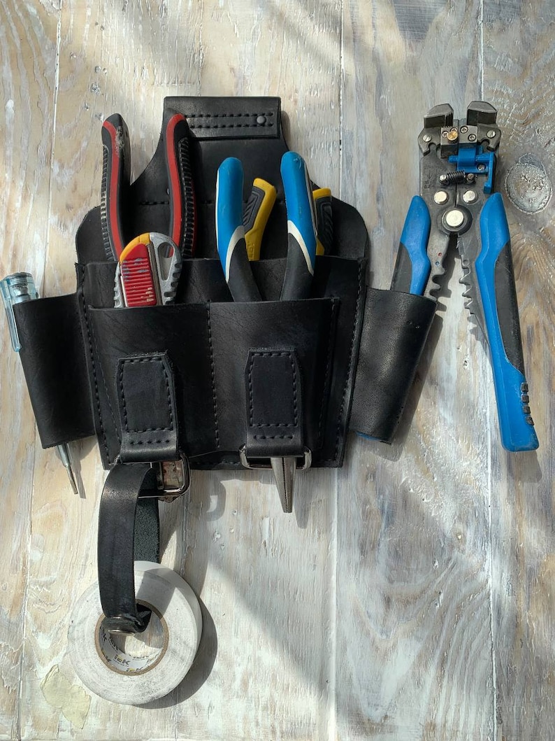 Peut inclure: Une ceinture &agrave; outils en cuir noir avec plusieurs poches, contenant divers outils tels que des pinces, des d&eacute;nudeurs de fils et un tournevis. La ceinture est accroch&eacute;e &agrave; une surface en bois.