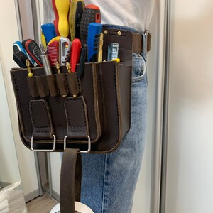 May include: A brown leather tool belt with multiple pockets and a loop for hanging a tool. The belt is worn by a person and has a variety of tools in the pockets, including screwdrivers, pliers, and a utility knife. A roll of tape is hanging from the loop on the belt.