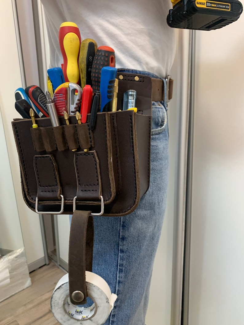 May include: A brown leather tool belt with multiple pockets and a loop for hanging a tool. The belt is worn by a person and has a variety of tools in the pockets, including screwdrivers, pliers, and a utility knife. A roll of tape is hanging from the loop on the belt.